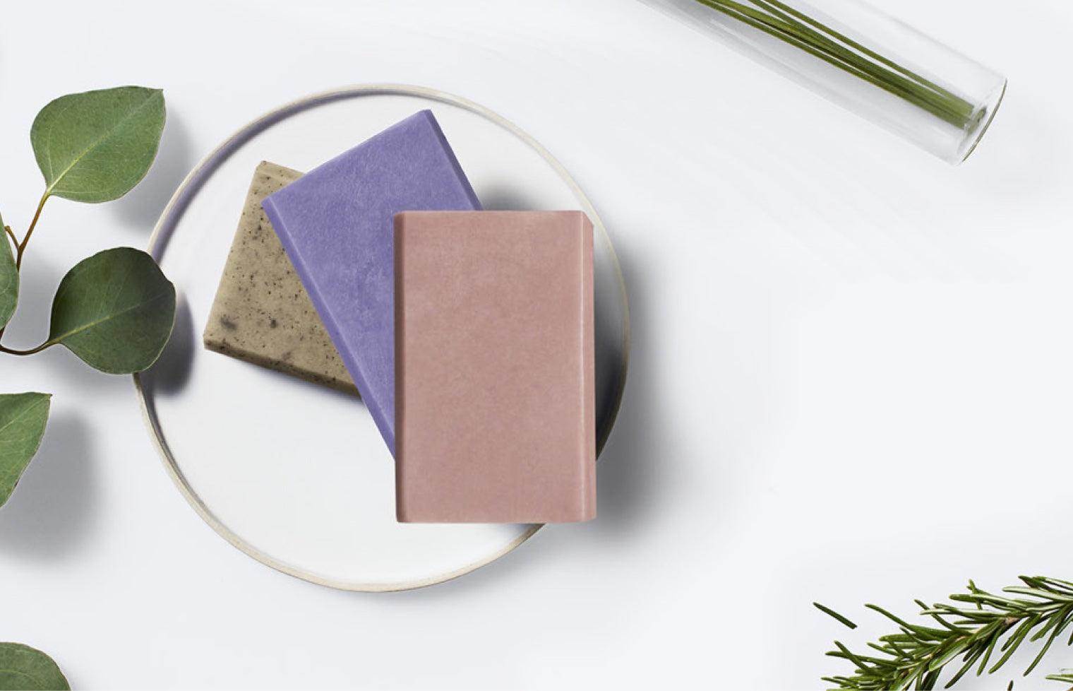 Three bars of soaps positioned on a table surrounded by herbs