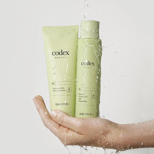 Codex Beauty Labs model's hand holding both Bia Exfoliating Wash and Bia Cleansing Oil. While water is raining down on the products and hand.