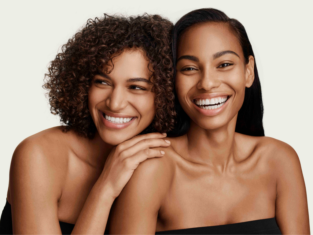 Two women smiling together in wrapped in black towels in front of a light beige background.