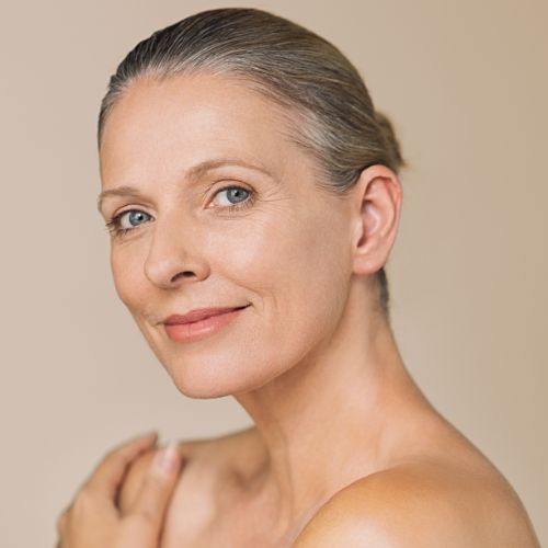A photograph featuring a woman in her middle age, holding her shoulders while standing against a brown background.
